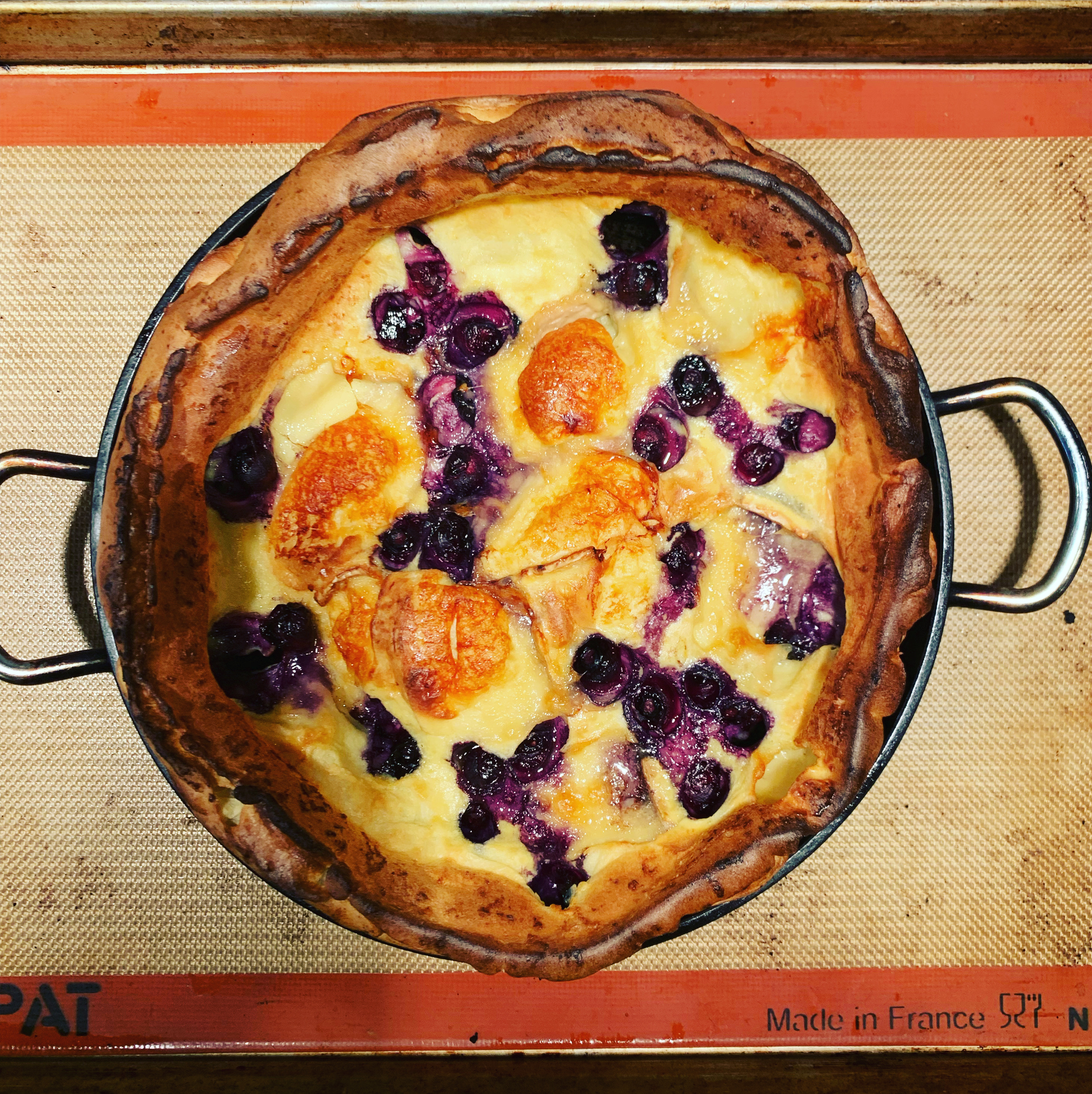 overhead photo of a baked Dutch baby with blueberries and cheese on a baking sheet with silicone mat on top